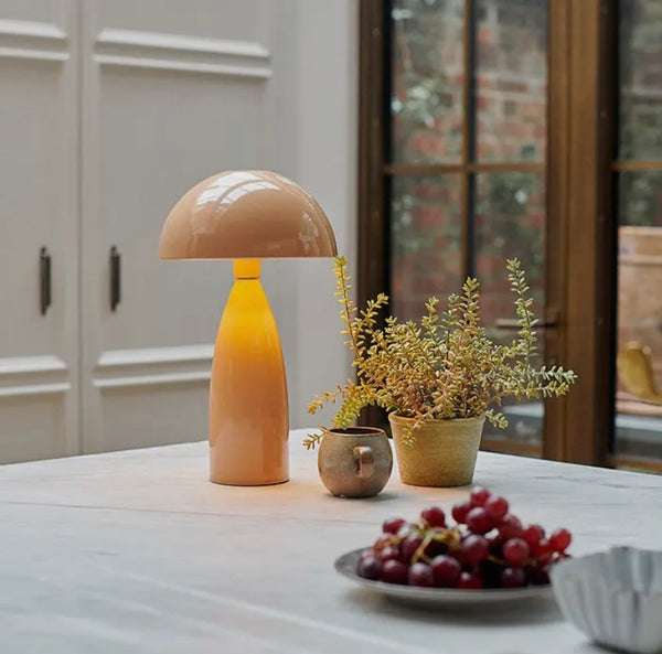 Blush domed table lamp emitting a light glow as it sits on a table next to a mug and a vase with some shrubs 