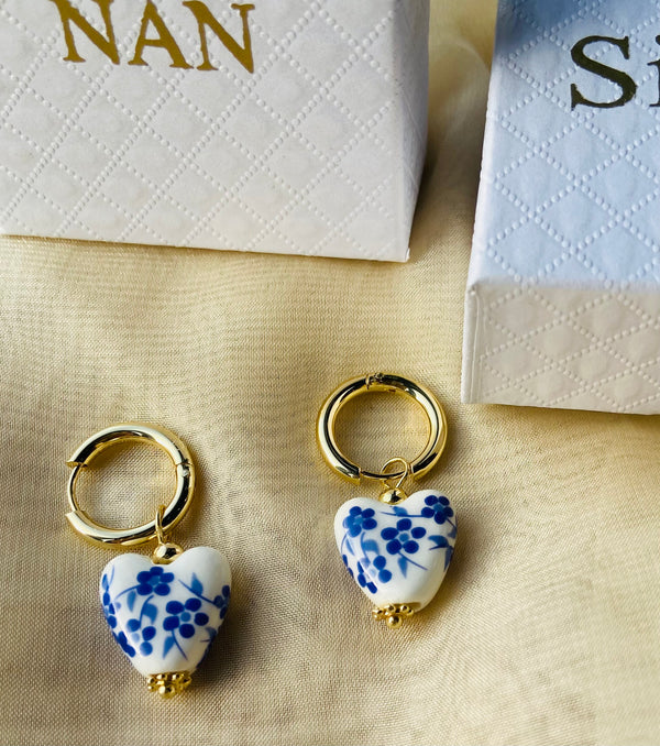 a pair of blue and white ceramic hearts on a gold hoop.  in the background is a white gift box with the word nan written in gold on the lid