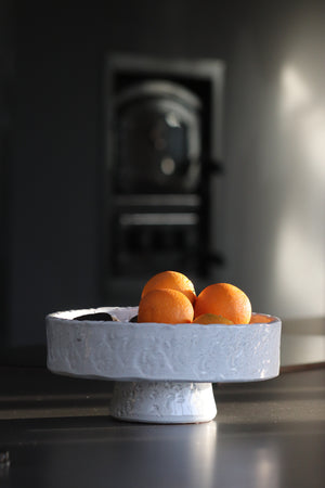 white textured bowl made from terracotta sits on black table with pizza oven in background.  Bowl contains fruit