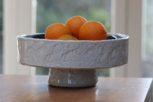 white textured bowl made from terracotta with oranges in it.  Sits on brown table with window in the background