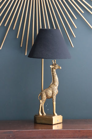 gold coloured giraffe table lamp with black shade on table with gold mirror prongs behind it