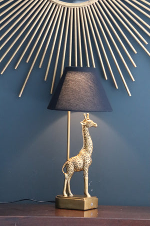 gold coloured giraffe table lamp with black shade on table with gold mirror prongs behind it.  The lampshade is on bathing the giraffe in a gold light