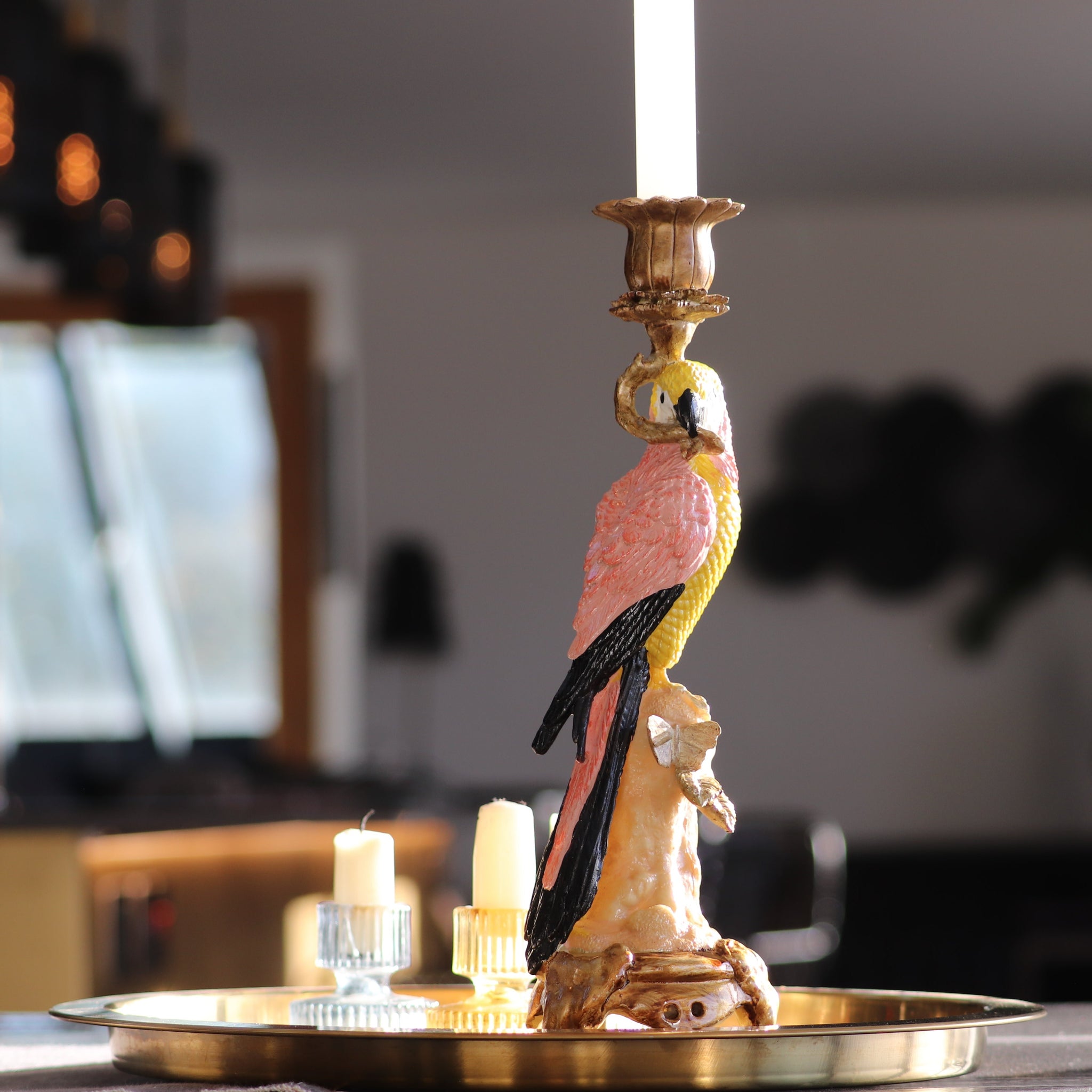 Wooden Parrot Colourful Candlestick Holder sat on a gold tray with two short glass candlestick holders - artwork in background