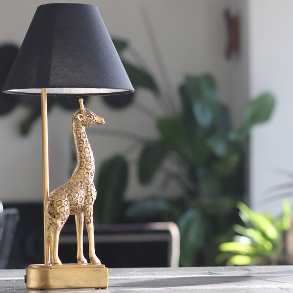 gold coloured giraffe table lamp with black shade on table with sunlight in background