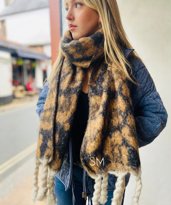 A blonde model stands in a doorway wearing a large  personalised blanket leopard print scarf with the initials SM embroidered in the corner