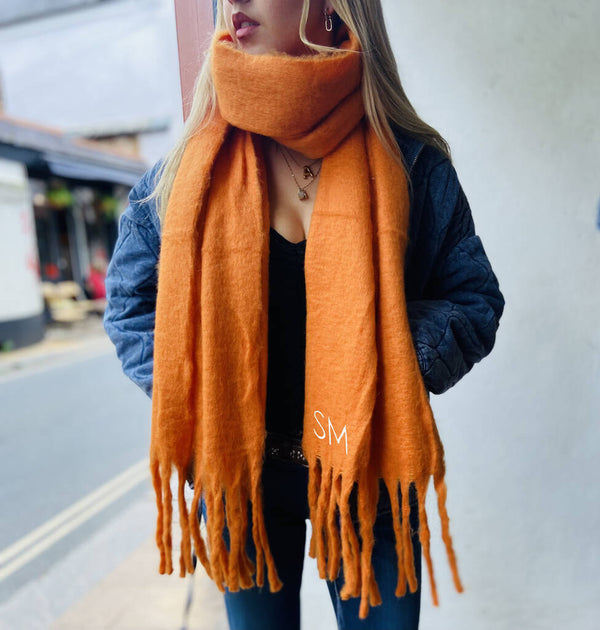 Model wears orange chunky personalised blanket scarf with the initials SM sewn into the bottom of the scarf just above the fringe