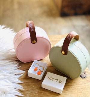 two personalised pink and sage green jewellery case holders, with lettering on the handle.  A gift box with sister embossed on it sits in front of them with a pair of orange drop gold hoop earrings