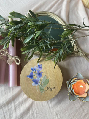 Round wooden personalised jewellery box in forefront with blue cornflower flower picture and the word Megan engraved onto it.  Beside the box is a pink candle wrapped in a string bow with a heart engraved with love attached