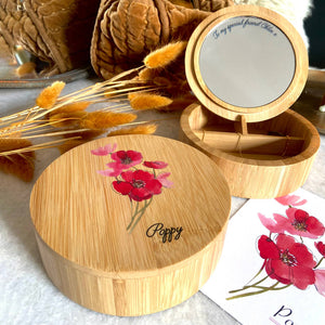 Round wooden personalised jewellery box in forefront with poppy flower picture and the word poppy engraved onto it.  In the background  the box is open showing a mirror and a card with the details of the birth flower