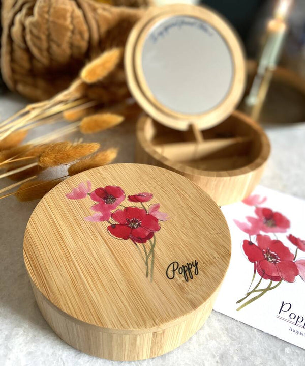 Round wooden personalised jewellery box in forefront with poppy flower picture and the word poppy engraved onto it.  In the background  the box is open showing a mirror and a card with the details of the birth flower