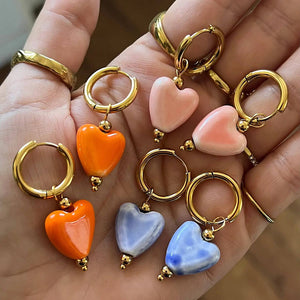 In the palm of a hand 3 sets of heart enamel earrings.  the heart dangles off a gold hoop earring. Colours are blue, orange and pink